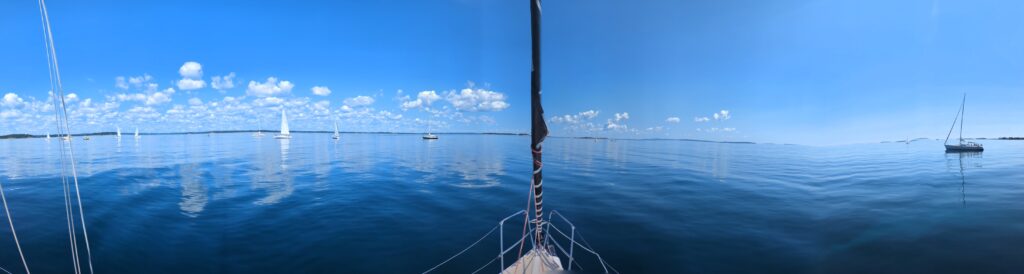 A panorama off of the bow of JahMon. Multiple sailboats are floating in calm waters with their sails down awaiting the winds to pick up.