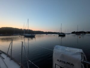 A view of the sunrise over the mooring field taken from the cockpit of SV Isola Naida. A few sailboats sit silhouetted against a cotton-candy sky reflected in the still ocean water.
